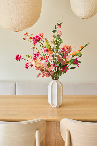 Op een lichte houten tafel staat een witte vaas met een boeket „Zijden kunstbloemen Wild Peach”, samengesteld uit zomerse bloemen. Twee lichte houten stoelen en grote papieren hanglampen maken het minimalistische, gezellige interieur compleet.