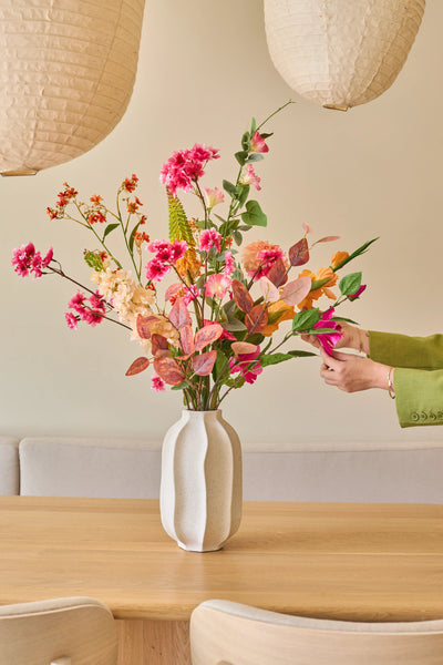 Iemand met een groene mouw schikt het boeket ‘Zijden kunstbloemen Wild Peach’, met kleurrijke zomerse bloemen, in een witte vaas op een lichte houten tafel, met papieren lantaarns en een beige bank op de achtergrond.