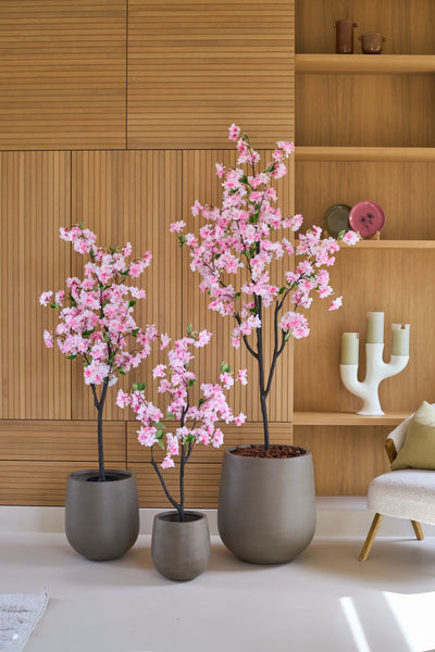 Drie Kunst Kersenbloesem-bomen van 150 cm met roze bloesems staan voor een houten lambrisering, naast planken die versierd zijn met decoraties en een moderne witte kandelaar.