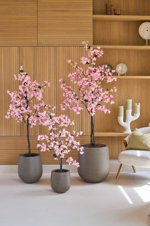 Drie Kunstplant-bougainvilles van 180 cm, roze van kleur en met roze bloemen, staan voor houten wandplanken, naast een moderne witte stoel met een kussen.