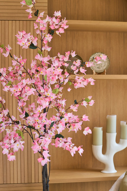An artificial bougainvillea plant of 180 cm, pink in color with pink flowers, stands in front of wooden shelves on which a decorative plate and a white candlestick with three green candles stand.