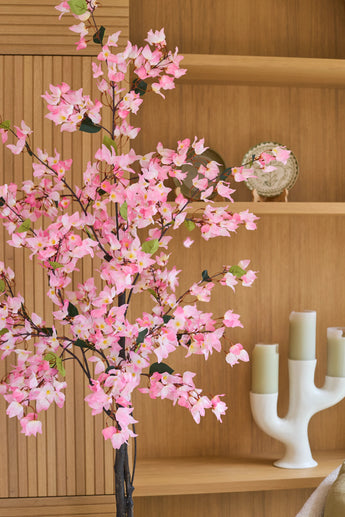 Voor de houten planken, waarop een bord en een witte kandelaar met drie groene kaarsen staan uitgestald, staat een kunstplant Bougainvillea van 120 cm in roze.
