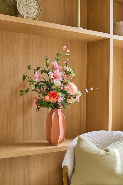 On a wooden shelf next to a cozy chair with a green cushion is a pink vase with the 'Yasmine's Choice' bouquet of silk artificial flowers, creating a warm and inviting interior image.