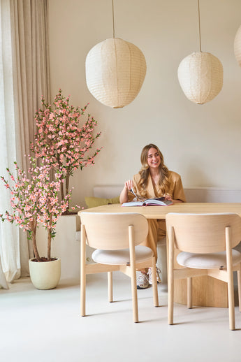A woman with long blond hair is sitting at a light wooden table, reading a book and smiling. Next to her is the Artificial Cherry Blossom Plant 150 cm pink, whose pink blossoms provide a lively accent. Hanging lamps and minimalist decor create a light, cozy atmosphere.