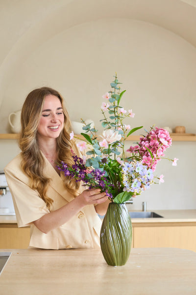 Een vrouw met lang, golvend haar schikt een boeket ‘Lavender Dreams’ van Zijden kunstbloemen in een groene vaas in haar moderne keuken, terwijl ze glimlachend aan het werk is. Het levendige boeket van zijden bloemen brengt tijdloze schoonheid in haar huis.