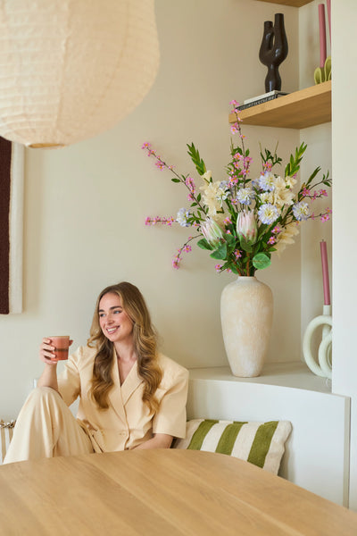 A woman in a cream-colored suit sits on a bench with a cup in her hand and a smile on her face. Behind her is a large vase with the artificial silk flower bouquet 'Lots of Love XL', consisting of pink, white and yellow flowers, with shelves in the background on which decorative objects are displayed. The setting looks cozy and modern.