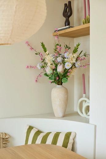 On a white shelf stands a beige vase with the colorful bouquet of artificial silk flowers, 'Lots of Love XL', next to pink candles and decorative objects. There is a green and white striped cushion on a chair under the shelf.