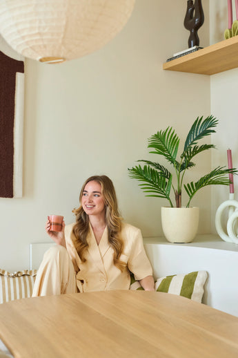 Een glimlachende vrouw met lang blond haar zit in een moderne, lichte kamer met een roze kopje in haar hand. Om haar heen staan groene planten, waaronder de weelderige Kunstplant Phoenix van 85 cm, evenals houten planken en decoratieve voorwerpen.