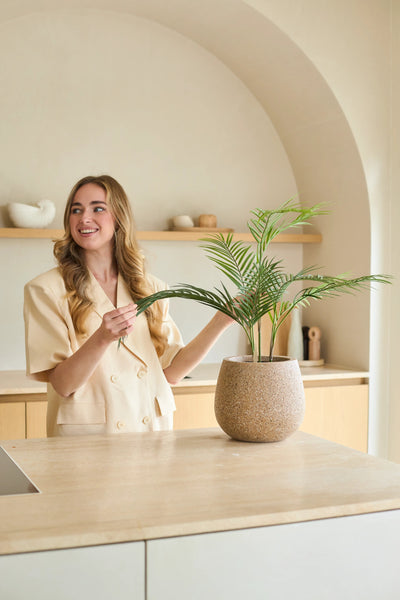 Een vrouw met lang blond haar glimlacht terwijl ze een Kunstpalm Chamaedorea van 48 cm in een pot verzorgt in een moderne, in neutrale tinten gehouden keuken met een minimalistische inrichting.