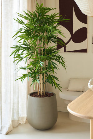 De Kunstplant Bamboe 150 cm, met weelderige groene bladeren, staat binnenshuis bij een raam met vitrages, naast een beige bankje met een kussen en abstracte wandkunst in een moderne kamer die is ingericht in neutrale tinten.