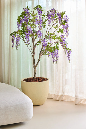 The Artificial Wisteria 210 cm plant, with its overhanging bunches of purple flowers, is placed indoors next to translucent beige curtains and a light-colored sofa.