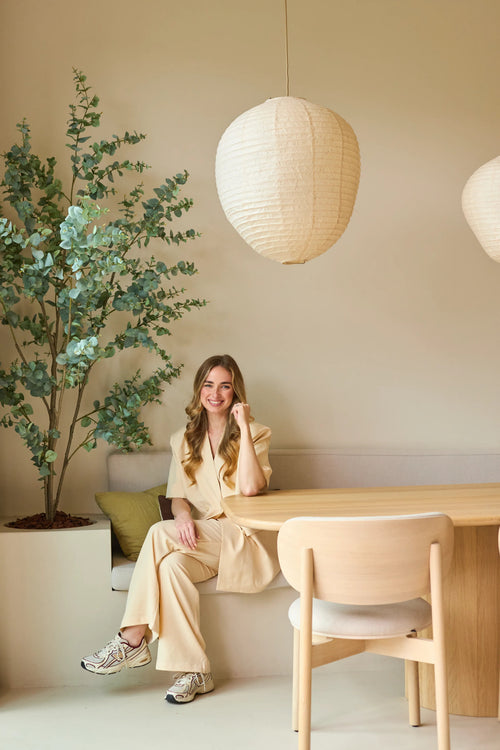 A woman in a beige suit sits smiling at a wooden table in a modern, minimalist room with light walls, paper pendant lamps hanging from the ceiling and a 180 cm Artificial Eucalyptus plant that adds some greenery to the room.
