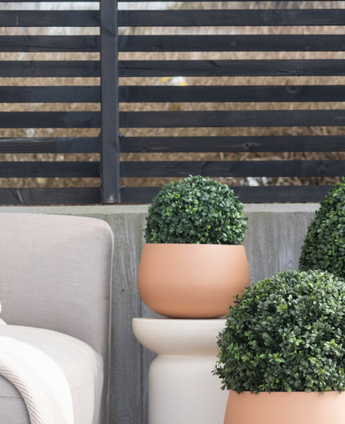 Three Artificial Plant Boxwood Roman Style UV D30cm round topiaries are on a patio near a beige outdoor chair, against a modern black slatted fence and bare trees.
