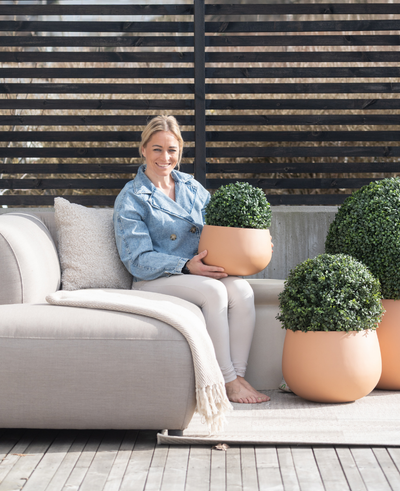 A woman sits barefoot on a light-colored outdoor bench, smiles and holds an Artificial Plant Boxwood Roman Style UV D30cm. Two similar artificial plants stand nearby, with wooden slatted fencing and patio decor in the background.