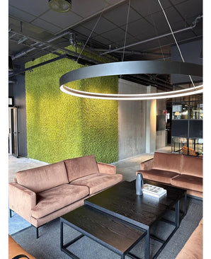 Een moderne zithoek met beige banken, zwarte salontafels, een grote ronde plafondlamp en op de achtergrond een groene muur, gecreëerd met Kunsthaag Nordic Mos 50x50 cm UV, die een luxe interieur en een eigentijds industrieel ontwerp uitstraalt.