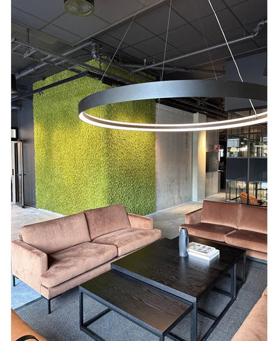 Een moderne zithoek met beige banken, zwarte salontafels, een grote ronde plafondlamp en op de achtergrond een groene muur, gecreëerd met Kunsthaag Nordic Mos 50x50 cm UV, die een luxe interieur en een eigentijds industrieel ontwerp uitstraalt.