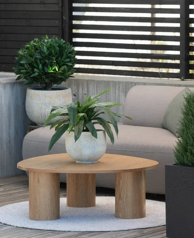 On a small rug next to a gray sofa on a terrace is a wooden coffee table with an artificial plant Bird's Deluxe of 60 cm on top, with a few potted plants in the background.