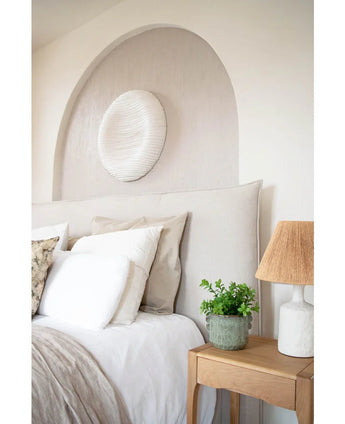 Een gezellige slaapkamer met een beige hoofdeinde, neutraal beddengoed en kussens, een houten nachtkastje met daarop de Kunstplant Eucalyptus 27 cm, een lamp met een getextureerde kap en ronde witte muurkunst boven het bed in een gewelfde nis.