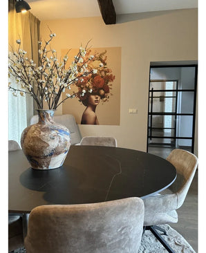 A modern dining area with a round black table, beige chairs and a large ceramic vase with Artificial Japanese Cherry Blossom 94 cm white. A mural of a woman with flowers in her hair decorates the background.