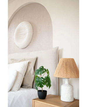 Een keurig opgemaakt bed met wit linnengoed staat naast een houten nachtkastje met daarop de Kunst Monstera 40 cm in sierpot zwart en een witte lamp met een geweven kap. Aan de muur hangt een zachte boog en een rond kunstwerk met structuur.