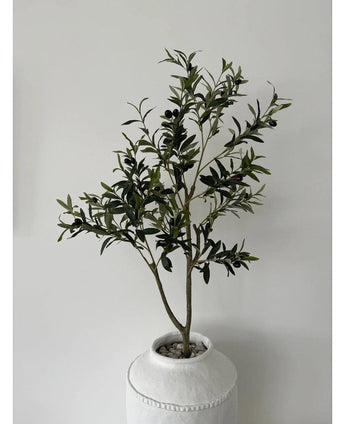 The Artificial Olive Tree 120 cm has green leaves and black olives in a white pot with structure and pebbles, making it a beautiful decorative element against a plain light gray wall.