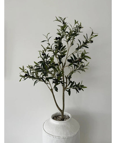 The Artificial Olive Tree 120 cm has green leaves and black olives in a white pot with structure and pebbles, making it a beautiful decorative element against a plain light gray wall.