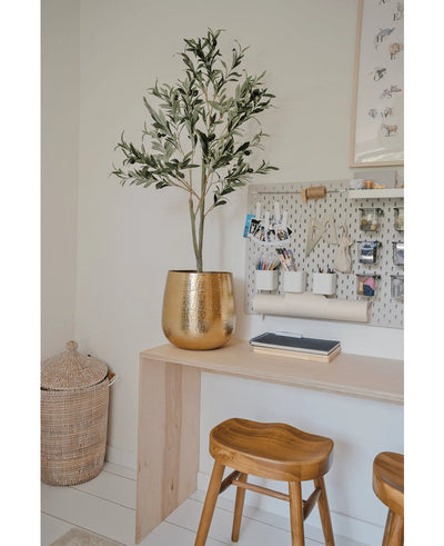 A modern workplace with a wooden desk, two stools, a large gold planter with the Artificial Olive Tree 120 cm for a Mediterranean flair, a washboard with office supplies and a wicker basket on the floor as a decorative detail.