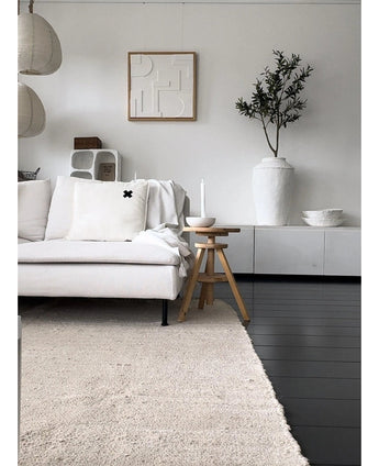 A minimalist living room with a light-colored sofa, neutral rug, white sideboard, Artificial Olive Tree 120 cm as a decorative element, abstract wall art and wooden stools - all in a soft Mediterranean atmosphere with neutral tones.