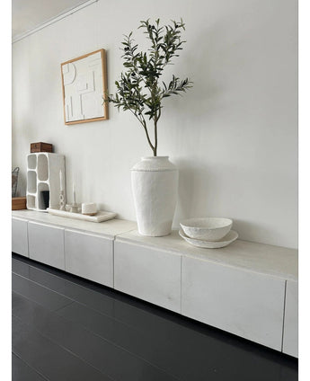 A minimalist room with a white sideboard, the Artificial Olive Tree 120 cm as a decorative element, decorative white bowls, a candle and abstract wall art. Dark wood floors and modern accents create a serene Mediterranean atmosphere.