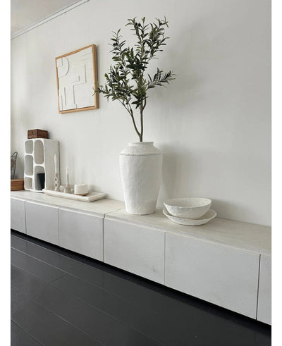 A minimalist room with a white sideboard, the Artificial Olive Tree 120 cm as a decorative element, decorative white bowls, a candle and abstract wall art. Dark wood floors and modern accents create a serene Mediterranean atmosphere.