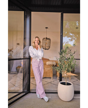 Een vrouw in een witte blouse en een lavendelblauwe broek glimlacht bij een grote glazen deur. De Kunst Olijfboom 120 cm brengt een mediterrane sfeer in de gezellige woonkamer, met een rieten bank en licht decor op de achtergrond.