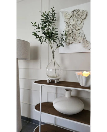 A minimalist console table shows a clear vase with the Artificial Olive Branch 109 cm, a round white ceramic vase and a white bowl with LED candles. Structured white wall art hangs above it and a floor lamp sits nearby.