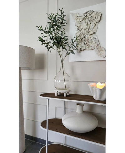Een minimalistische consoletafel toont een heldere vaas met de Kunst Olijftak 109 cm, een ronde vaas van wit keramiek en een witte schaal met LED-kaarsen. Erboven hangt gestructureerde witte wandkunst en vlakbij staat een staande lamp.