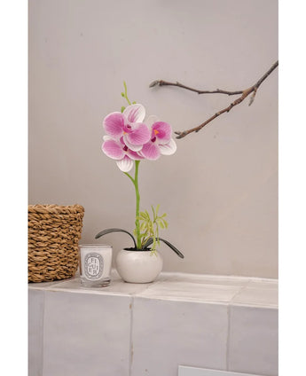 The Artificial Orchid 28 cm white/pink in pot stands on a tiled surface next to a woven basket and a white Diptyque scented candle, against a light wall with a bare branch above it.