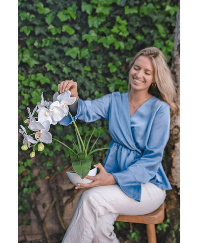 Een glimlachende vrouw in een lichtblauwe blouse en witte broek zit op een krukje, houdt de Kunst Orchidee 50 cm wit in pot vast en raakt voorzichtig de bloemen aan. Ze zit tegen een achtergrond van groen gebladerte.