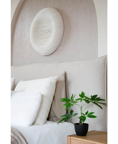 Een keurig opgemaakt bed met wit beddengoed staat naast een houten nachtkastje met de Kunst Pachira 30 cm plant. Boven het bed siert een wit gestructureerd cirkelvormig wandschilderij de lichtgekleurde muur.