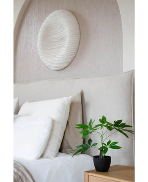 Een keurig opgemaakt bed met wit beddengoed staat naast een houten nachtkastje met de Kunst Pachira 30 cm plant. Boven het bed siert een wit gestructureerd cirkelvormig wandschilderij de lichtgekleurde muur.