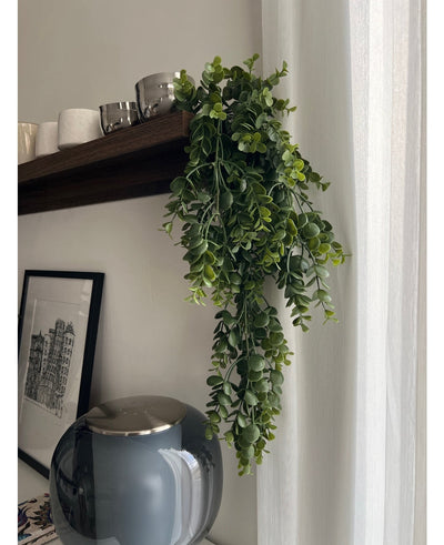 De Kunst hangplant Pannenkoek 61 cm hangt aan een houten plank boven een grijze glazen vaas, met kopjes en schalen ernaast. Een ingelijste zwart-wit stadsschets leunt tegen de muur bij een wit gordijn.