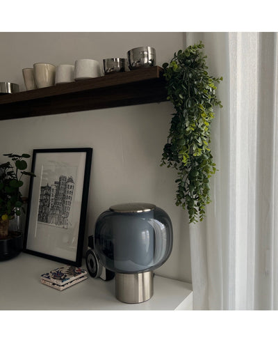Een modern tafelblad met een lamp van donker glas, een ingelijste schets en onderzetters ligt onder een houten plank met kopjes en mokken. De Kunst hangplant Pannenkoek 61 cm hangt aan de plank, terwijl transparante gordijnen zacht natuurlijk licht binnenlaten.