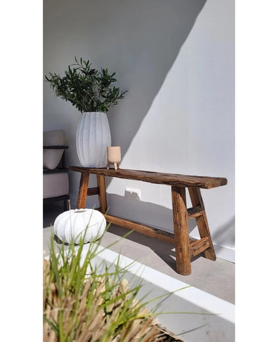 A rustic wooden bench stands against a white wall, next to a tall white vase with Artificial olive leaf 90 cm UV for a Mediterranean atmosphere. Nearby are a round white ottoman and a small candlestick, with sunlight casting soft shadows.
