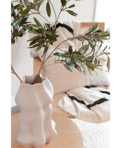 A white sculpted vase holds Art olive leaf 90 cm UV with green leaves on a light wooden table. Behind it, a beige chair and two cream-colored cushions with striking black stripes enhance the Mediterranean atmosphere.