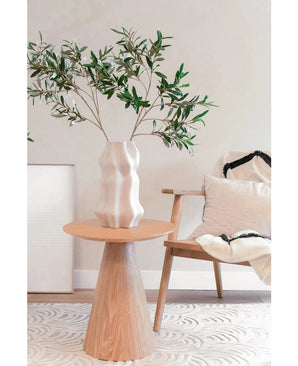 In een moderne, minimalistische leefruimte met mediterrane inrichting staat een gebeeldhouwde witte vaas met het Kunst olijfblad 90 cm brandvertragend op een ronde houten tafel, naast een lichte houten stoel met witte en zwarte kussens.