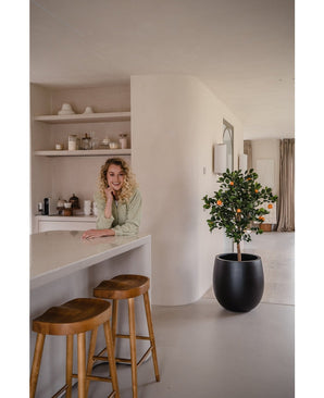 A woman with curly blond hair smiles, leaning on a counter. Next to her, two wooden stools and an artificial orange tree 120 cm give a fresh atmosphere to the modern, bright kitchen with shelves and plates behind it.