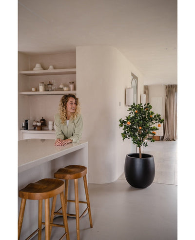 Een vrouw met krullend blond haar glimlacht achter een aanrecht met twee houten krukjes. De Kunst sinaasappelboom 120 cm in een pot voegt charme toe aan de moderne, minimalistische ruimte die is ingericht in neutrale tinten.