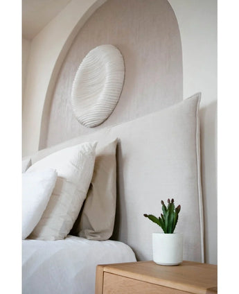 A modern bedroom has a beige upholstered headboard, white pillows, an artificial succulent 20 cm on a wooden bedside table and round textured wall art above the bed.
