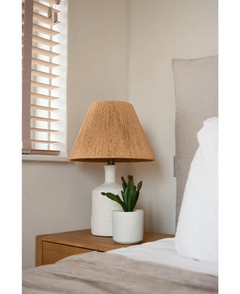 Een nachtkastje met een witte lamp met een geweven beige kap en de Kunst vetplant 20 cm staat naast een bed met wit en beige beddengoed, bij een raam met houten jaloezieën.