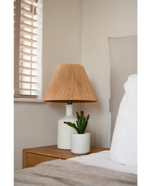 A bedside table with a white lamp with a woven beige shade and the Artificial succulent plant 20 cm stands next to a bed with white and beige bedding, near a window with wooden blinds.