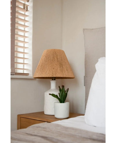 A bedside table with a white lamp with a woven beige shade and the Artificial succulent plant 20 cm stands next to a bed with white and beige bedding, near a window with wooden blinds.