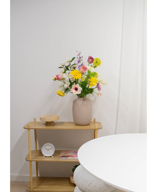 A light wooden shelf shows a beige vase with Artificial Ranunculus Real Touch Yellow 60cm flowers, a white clock and a wooden tray. Part of a round white table appears in the foreground against a plain white wall.