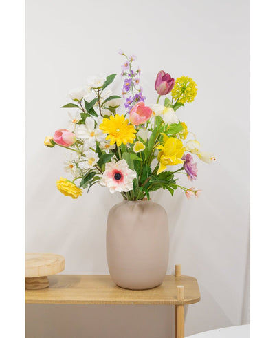 A beige vase with an Artificial Ranunculus Real Touch Yellow 60cm artificial flower stands on a light wooden table against a plain white wall.
